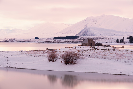 Winter in Tekapo, New Zealand.  Looking across the river to the Church of the Good Shepherd and mountains beyond, the whole landscape is covered in fresh snow. The soon-to-rise sun is casting a pink glow on the scene.