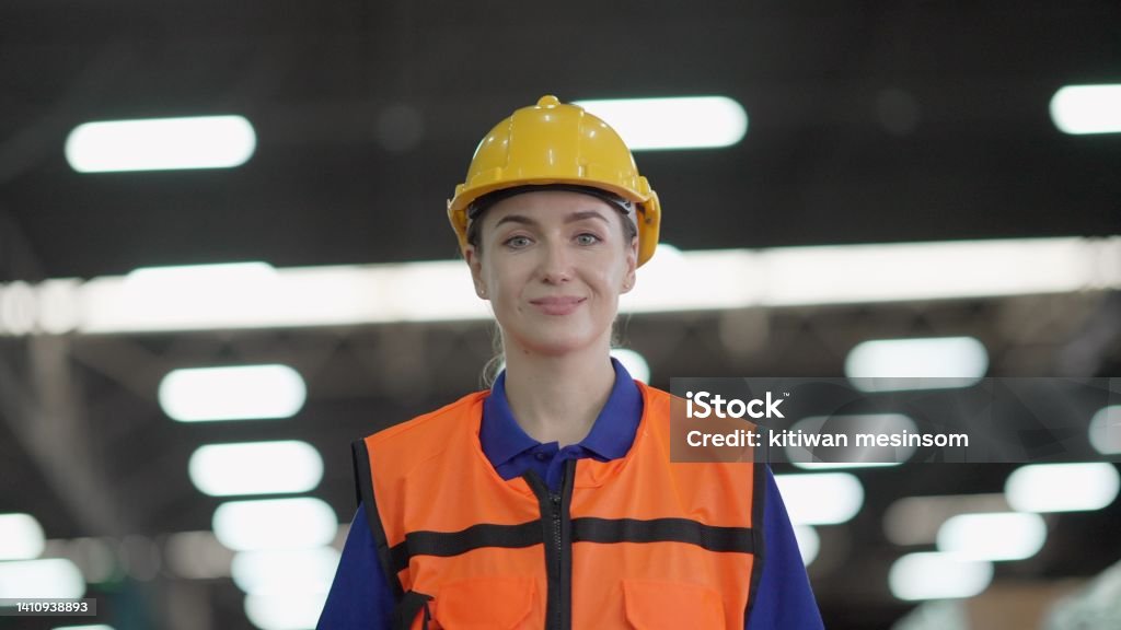 Professional female manager or worker wearing hard hat and safety vest, have confident and leadership, walking to work in the retail warehouse, smiling and looking at the camera. Employee working on product distribution logistics center. Adult Stock Photo