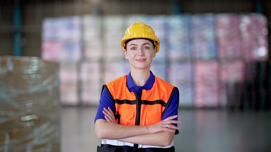 Professional female manager or worker wearing hard hat and safety vest, have confident and leadership, standing among the employees working in the retail warehouse, crossed arm, smiling and looking at the camera. Employee working on product distribution logistics center.