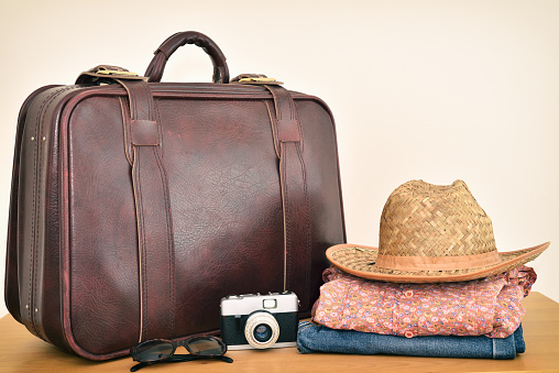 Vintage leather suitcase with straw hat, floral shirt, jeans, sunglasses and retro photo camera on the brown wooden table. Copy space. Travel baggage concept. Retro style photo filter added.