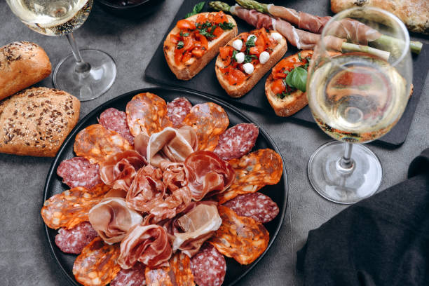 aperitivo de comida jamón serrano, salami, aceitunas. queso sobre tabla parmesano, pecorino, gorgonzola. bruschetta de tomate y albahaca. dos copas de vino blanco o prosecco - bruschetta cutting board italy olive oil fotografías e imágenes de stock