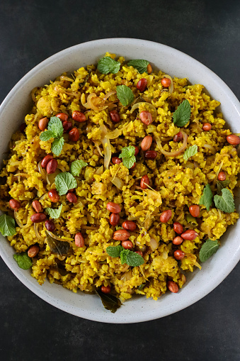 Stock photo showing close-up, elevated view of poha rice dish of spiced flattened rice with onion, green chillies, curry and coriander leaves, peanuts, mustard and cumin seeds, turmeric and lemon juice.
