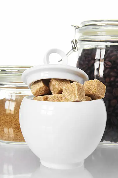 White sugarbowl with brown sugarcubes  and jars of coffee and sugar on white background