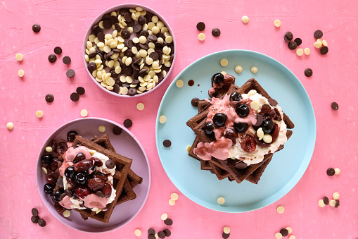 Stock photo showing close-up view of piles of homemade chocolate waffle squares topped with melting raspberry ice cream, blueberries, cherries, milk and white chocolate chips.