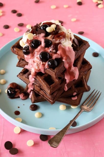 istock Close-up image of stack of homemade chocolate waffle squares topped with melting raspberry ice cream, blueberries and cherries, milk and white chocolate chips, light blue plate, fork, pink background, elevated view, focus on foreground 1410957370