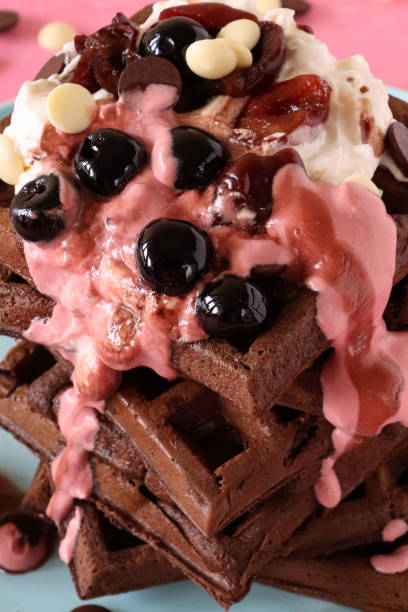 istock Full frame image of stack of homemade chocolate waffle squares topped with melting raspberry ice cream, blueberries and cherries, milk and white chocolate chips, light blue plate, pink background, elevated view, focus on foreground 1410957374