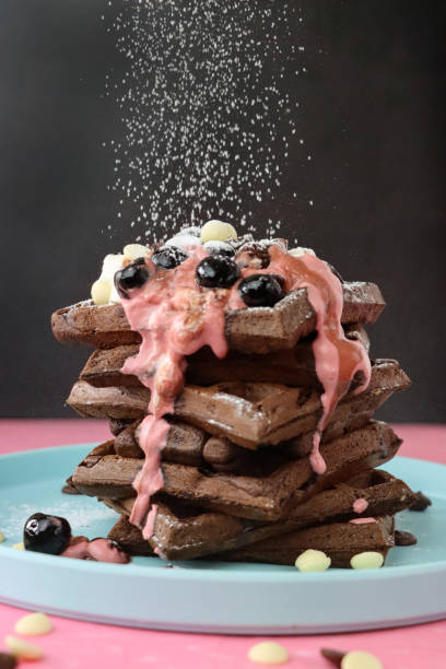 Image of icing sugar sifted onto stack of homemade chocolate waffle squares topped with melting raspberry ice cream, blueberries and cherries, milk and white chocolate chips, light blue plate, pink surface, black background, focus on foreground stock photo