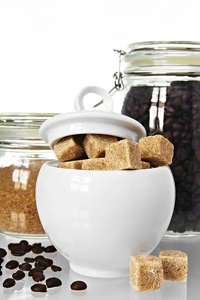 White sugarbowl with brown sugarcubes and jars of coffee and sugar on white background