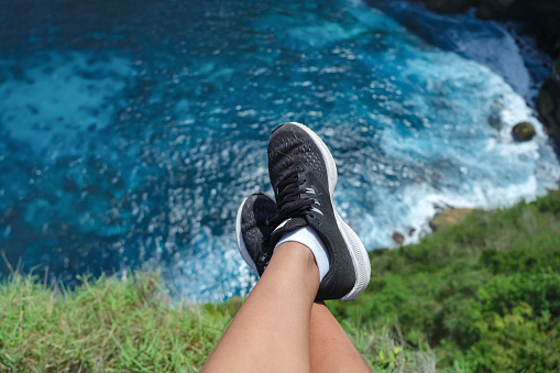 Close- up shot of unrecognizable hanging feet with shoes above a turquoise sea, sitting on the grass.