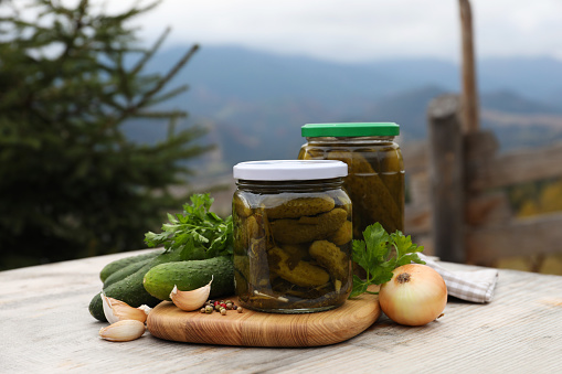 Jars of delicious pickled cucumbers and ingredients on wooden table in mountains