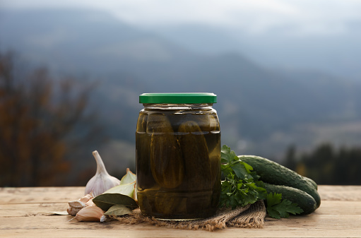 Jar of delicious pickled cucumbers and ingredients on wooden table in mountains