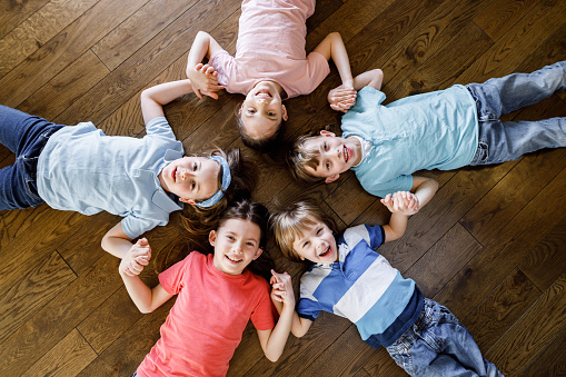 High angle view of happy kids having fun while holding hands and lying on the floor at home while looking at camera. Copy space.