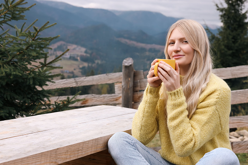 Young woman with mug of hot drink at wooden table in mountains