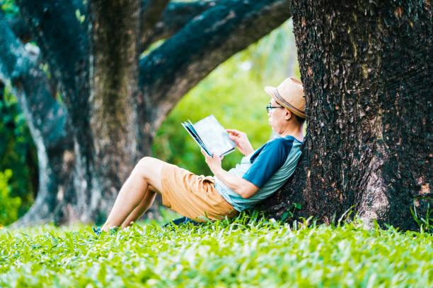 istock Asian man in casual clothes reading under a tree, sitting on the grass and relaxing with a book in the park with copy space 1410960136