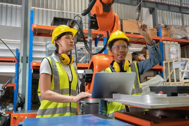 Women Engineer wearing helmet and hi visible vest using laptop computer working in factory welding robotic automation happy smiling with quality control and programming pass