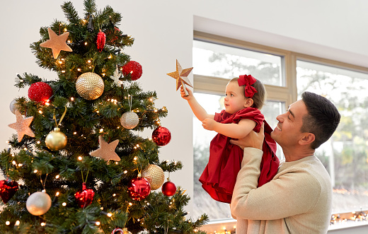 winter holidays and family concept - happy middle-aged father and baby daughter decorating christmas tree with star at home