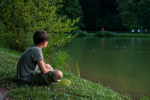 A boy in a green T-shirt sits thoughtfully on the green shore of the lake and looks thoughtfully.