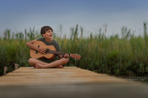 A boy play the guitar . He sits on the pier and plays a song on the guitar and sings. He shoots a guitar at the camera in the style of Johny Cash. In the background Behind him the river channel, the bank.