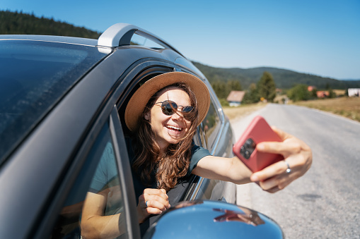 Happy young woman driver traveler in hat and sunglasses sitting behind the wheel of car taking selfie using smartphone