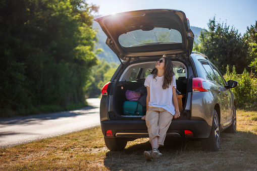 Happy young woman driver traveler in sunglasses standing near open car trunk. Road trips and summer holidays