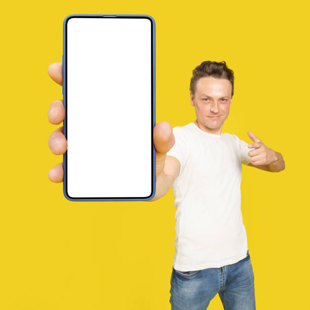 istock Handsome man holding huge smartphone with white screen pointing at white empty screen, wearing white t-shirt and jeans isolated on yellow background. Mobile app advertisement, great offer 1410970637