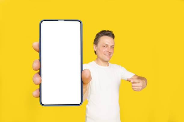 istock Huge smartphone in hand of handsome blond man pointing at white empty screen, wearing white t-shirt and jeans isolated on yellow background. Mobile app advertisement, great offer 1410970643
