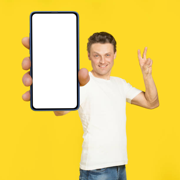 istock Gesturing V or victory handsome man holding huge smartphone with white screen wearing white t-shirt and jeans isolated on yellow background. Mobile app advertisement, great offer 1410970644