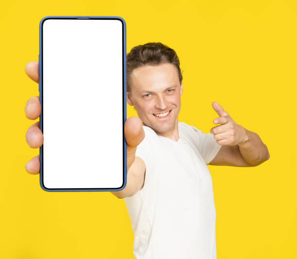 istock Holding huge smartphone with white screen handsome blond man pointing at white empty screen, wearing white t-shirt and jeans isolated on yellow background. Mobile app advertisement, great offer 1410970645