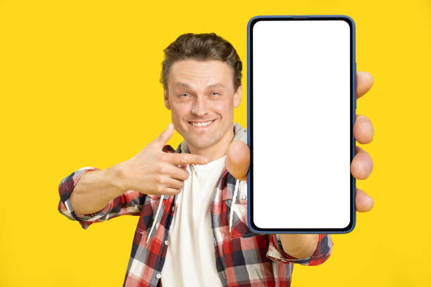 istock Pointing at giant, huge smartphone with white empty screen blond man, wearing red plaid shirt. Man with phone display mock up isolated on yellow background. Mobile app advertisement 1410970651