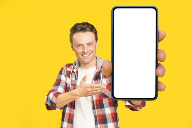 istock Introducing new app happy holding giant, huge smartphone with white screen blond man, wearing red plaid shirt. Man with phone display mock up isolated on yellow background. Mobile app advertisement 1410970652