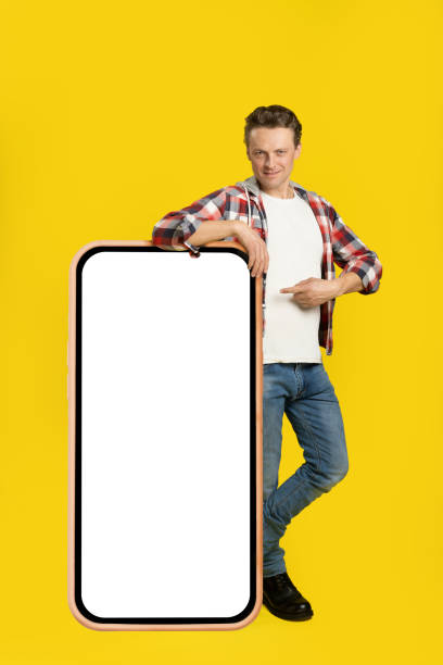 istock Handsome young man standing leaned on giant, huge smartphone, digital tablet with white, black screen pointing on dressed in plaid shirt and jeans isolated on yellow background. Free space mock up 1410970655