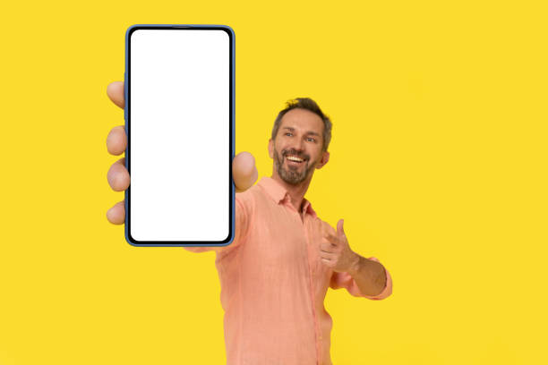 istock Middle aged grey haired man pointing at smartphone in hand amazed smiling looking at smartphone wearing peach shirt isolated on yellow. Mature fit man with phone, mobile app advertisement 1410975287