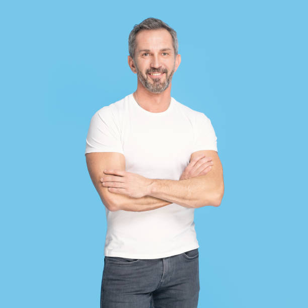 istock Handsome middle aged grey haired man standing with arms folded happy looking at camera wearing white t-shirt and jeans isolated on blue background. Mature fit man, healthy lifestyle concept 1410975578