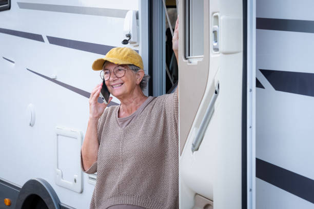 smiling senior woman in travel vacation leisure outside a camper van enjoying freedom and travel. attractive elderly caucasian lady in van life standing at the door of motor home talking on mobile phone - motor home mobile home camping senior adult imagens e fotografias de stock