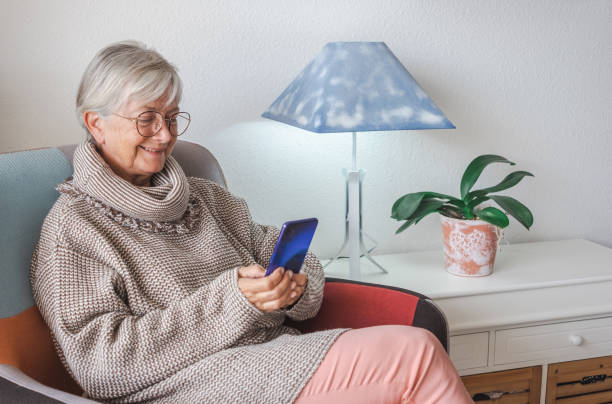 テクノロジーとソーシャルメディアを楽しむ携帯電話を使って自宅に座っている笑顔の美しいシニア女性 - 古い世代と新しい技術のコンセプト - old armchair women senior adult ストックフォトと画像