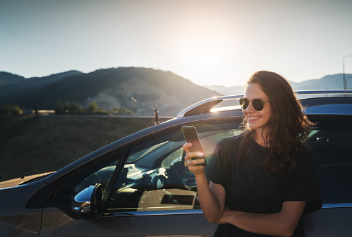 Young beautiful woman traveling by car in the mountains using smartphone at sunset