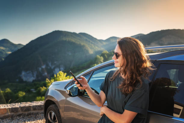 Young beautiful woman traveling by car in the mountains using smartphone at sunset, summer vacation and adventure stock photo