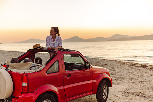 Woman in white shirt taking a break fro driving a car and enjoying the moment in sunset by the sea