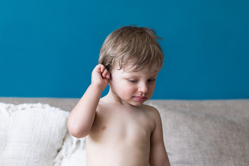 Cute little toddler boy rubs and scratches his ear with hand sitting by the sofa at home. Allergies or infection issues