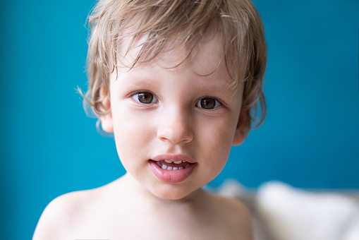 Portrait of little cute toddler boy looking at a camera sitting at home against blue background