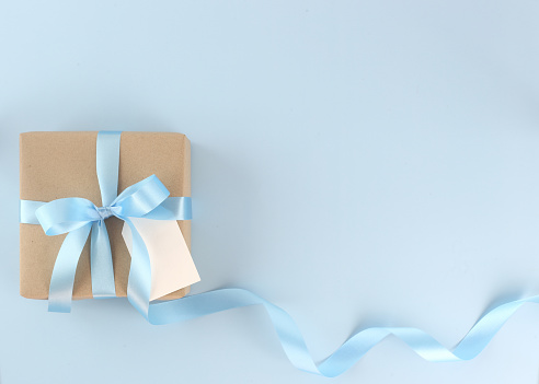Beautiful brown paper gift box with blue satin curly ribbon bow and blank card on pastel blue background. Flat lay Mother day, father day, valentines day, birthday concepts with copy space.