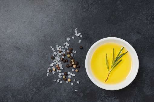 Olive oil in white bowl on black stone table with rosemary and salt and pepper. Top view, flatlay