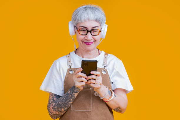 old senior asian woman wear apron hand choosing music playlist from smartphone relax casual lifestyle,asia tattoo woman wear headphone listen music carefree isolate yellow background studio shot stock photo