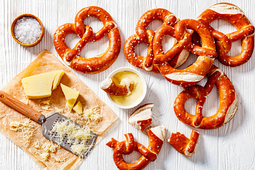 soft pretzels baked in the form of knot and sprinkled with salt on white wooden table with cheese sauce and grated cheese, horizontal view from above, flat lay