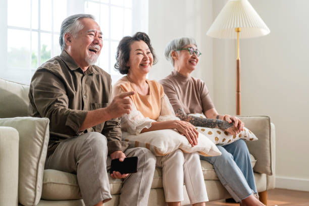 gruppe von asiatischen alten älteren männern und frauen, die wochenendzeit miteinander verbringen, sitzen auf der sofacouch und schauen tv-comedy-programm mit freudigem lachen lächelndes glück, ausdruck von senioren im pflegeheim - senior women electronics people remote control stock-fotos und bilder