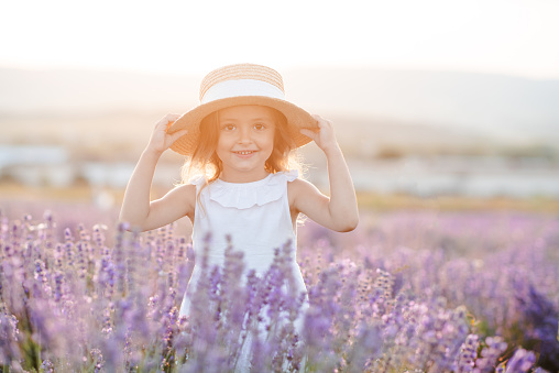 Happy smiling child girl 3-4 year old wear starw hat having fun in blooming lavender flower field over nature background outdoor. Childhood. Summer season.