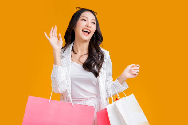 Asian happy female woman girl holds colourful shopping packages   standing on yellow background studio shot, Close up Portrait young beautiful attractive girl smiling looking at camera with bags stock photo