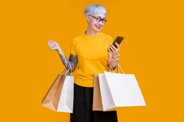 Asian happy senior female woman tattoo holds colourful shopping packages standing on yellow background studio shot, Close up Portrait old female hand hold smartphone enjoy shopping online cashless stock photo
