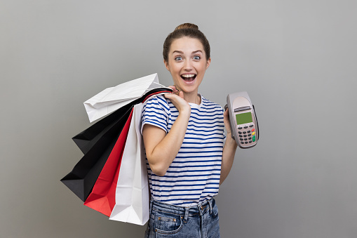 Portrait of smiling cheerful woman wearing striped T-shirt holding and showing payment terminal and shopping bags, expressing excitement. Indoor studio shot isolated on gray background.