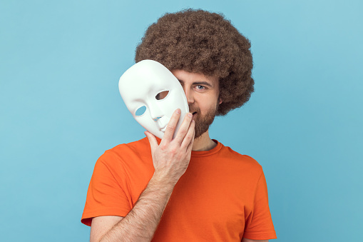Portrait of man with Afro hairstyle wearing orange T-shirt peeping out white face mask, hiding his real feelings, pretending to be another person. Indoor studio shot isolated on blue background.